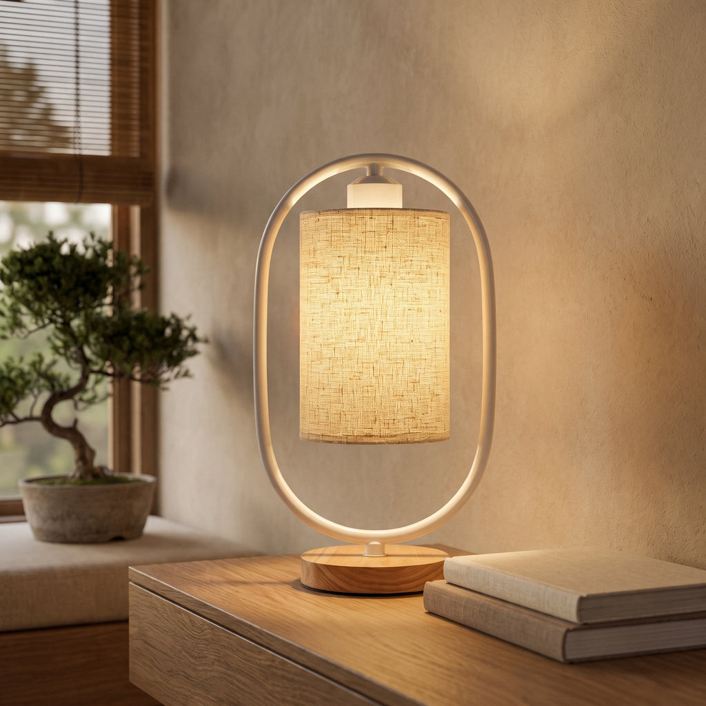 White oval frame table lamp with linen shade glowing on a wood surface by a window