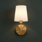 Single battery operated wall sconce in brushed brass with a white fabric shade, lit on a charcoal green wall