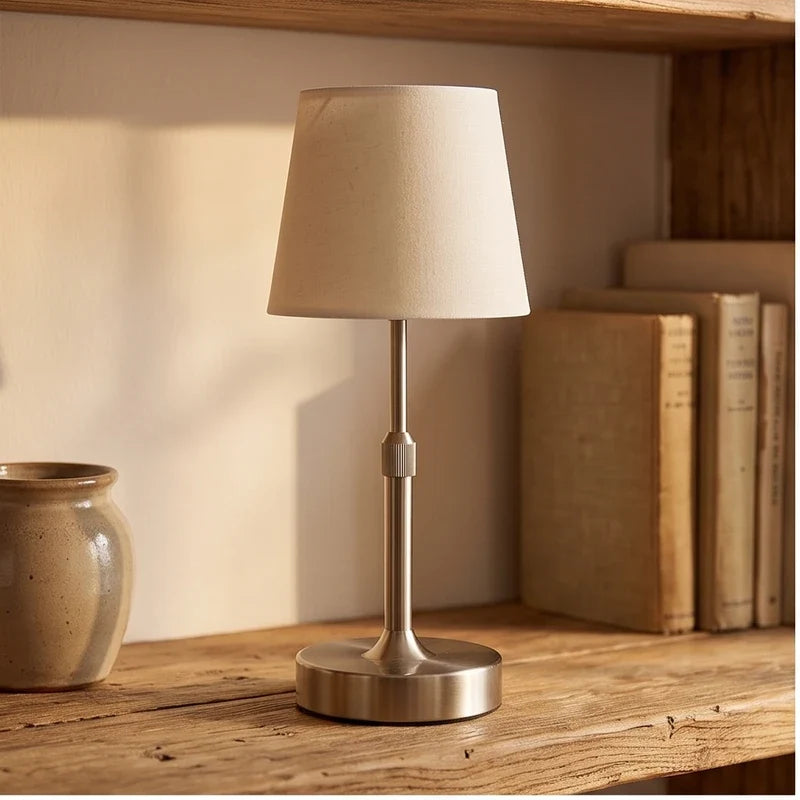 Silver cordless table lamp with neutral fabric shade on a wooden shelf