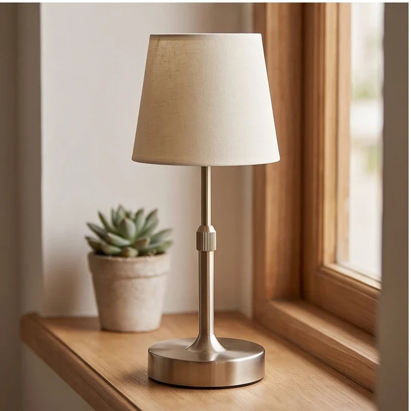 Gold cordless table lamp with cream fabric shade on a wooden window sill