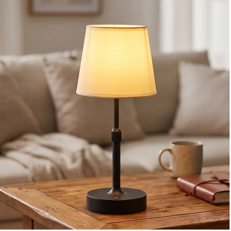 Black cordless table lamp glowing warm on a wooden table beside a mug and notebook