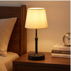 Black cordless table lamp glowing on a wooden nightstand beside a bed